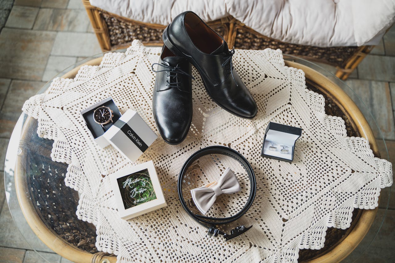 Flat lay of elegant men's accessories including shoes, belt, watch, and bow tie on a crochet tablecloth.